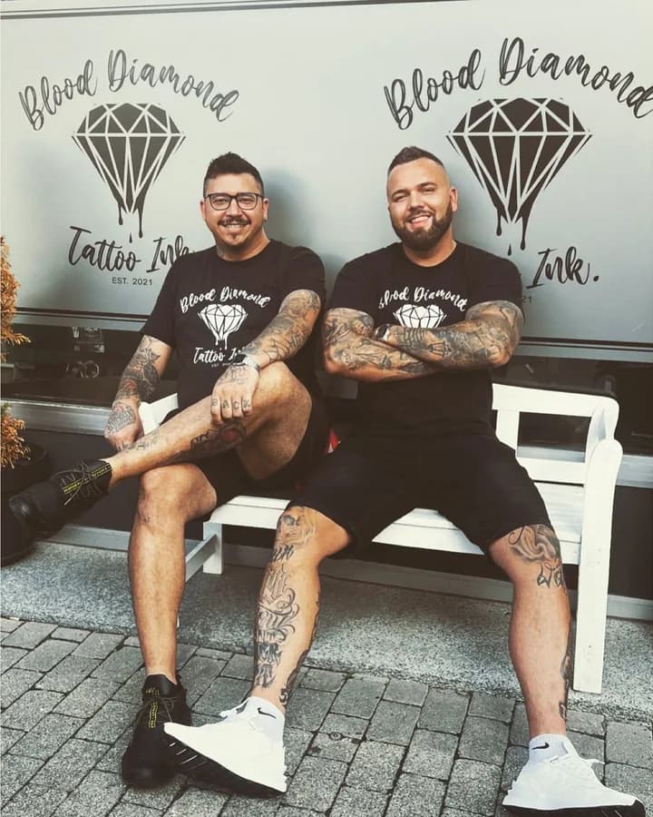 Zwei tätowierte Blood-Diamond-Teammitglieder sitzen lachend vor dem Studio-Schriftzug.